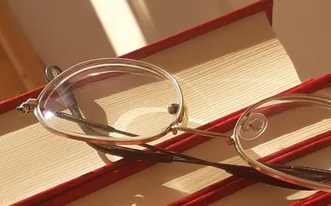 A pair of silver-rimmed eyeglasses resting on a stack of red books in warm light.