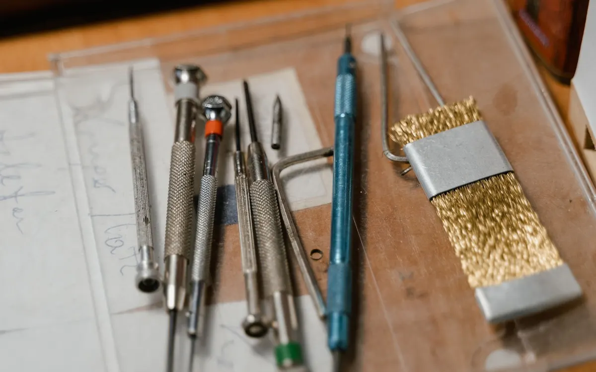 Metal precision tools, including screwdrivers, scribes and a brass brush, arranged on a wooden workbench beside technical drawings.