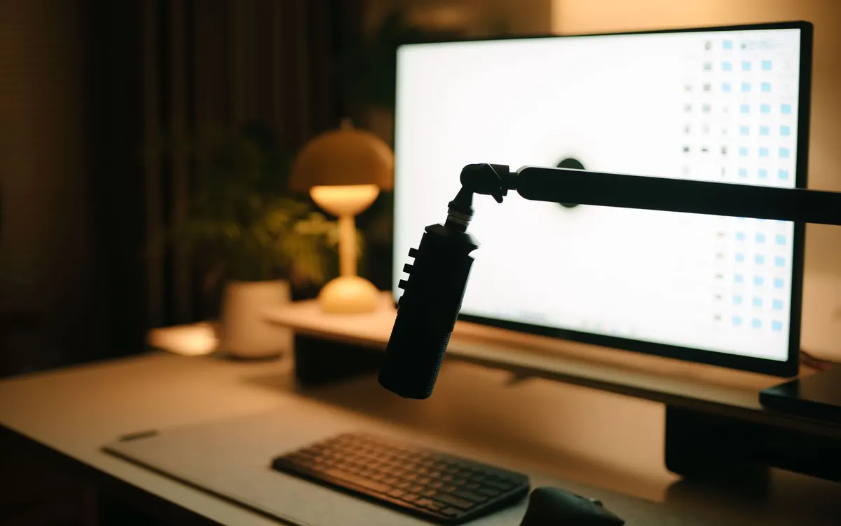 A black studio microphone on a boom arm beside a computer monitor, keyboard, lamp and plant on a wooden desk in warm light.