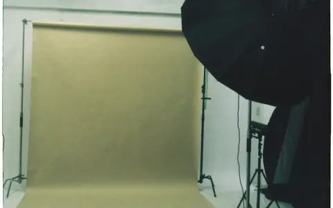 A photography studio with a beige paper backdrop mounted on stands and black lighting equipment with a softbox on a tripod.
