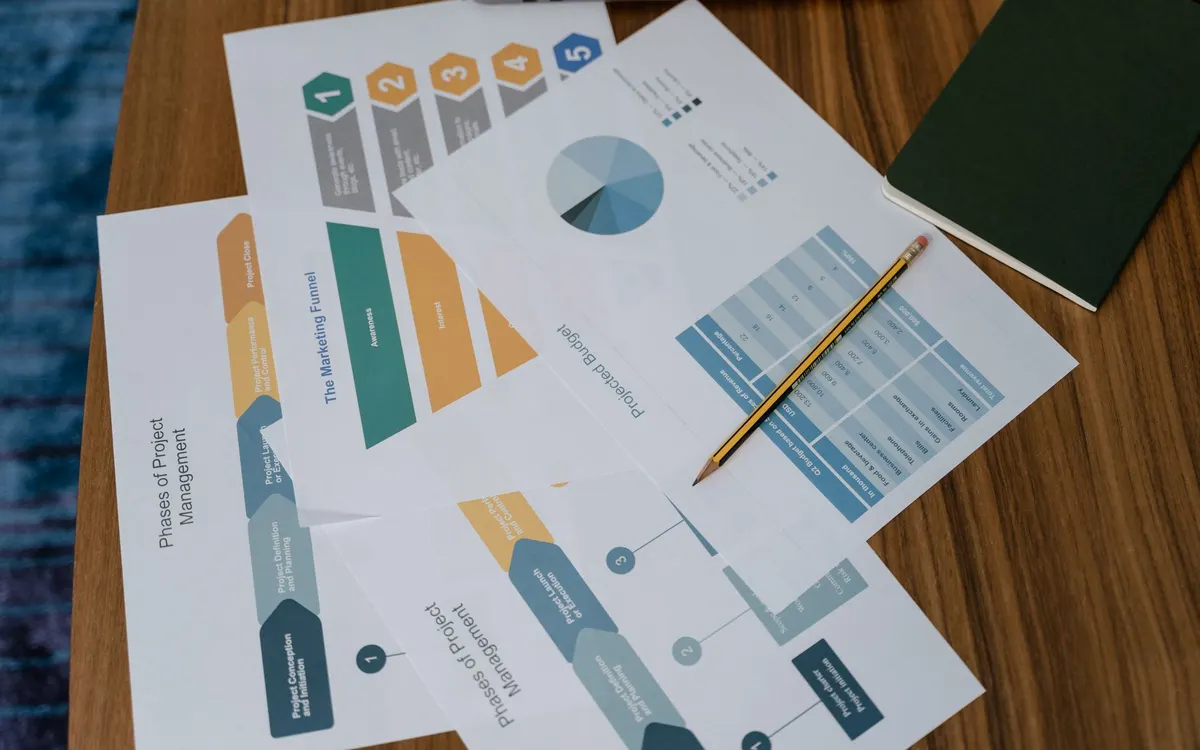 Printed infographic sheets showing charts and diagrams lie on a wooden desk with a pencil on top.