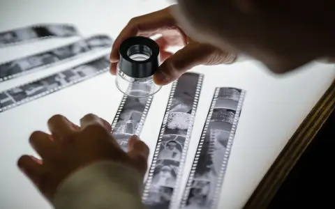 A hand holds a magnifier over strips of film negatives laid on a white lightbox surface.