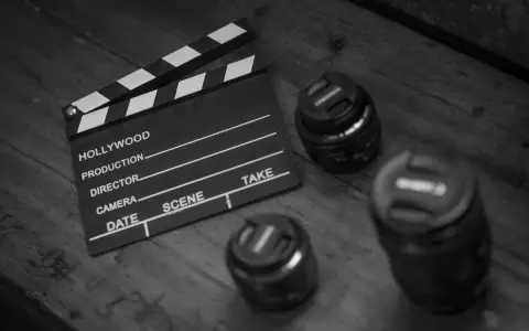 A monochrome film clapperboard rests on a wooden surface beside three camera lenses.
