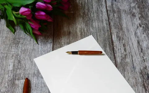 A fountain pen rests on blank white paper on a weathered wooden surface, with flowers at one corner.